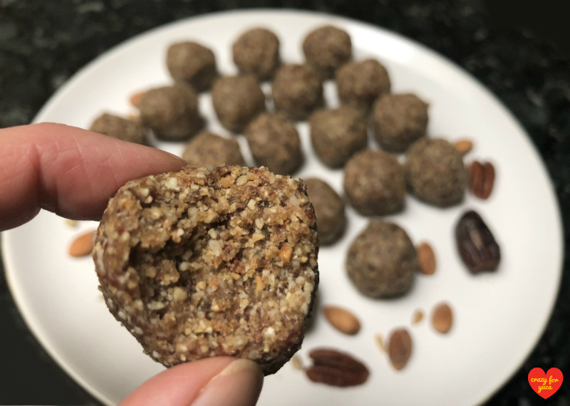 CrazyForYuca.com - Close up of one Paleo No-Bake Date Nut Ball with a bite taken out of it. Background is a white plate filled with Paleo No-Bake Date Nut Balls. 
