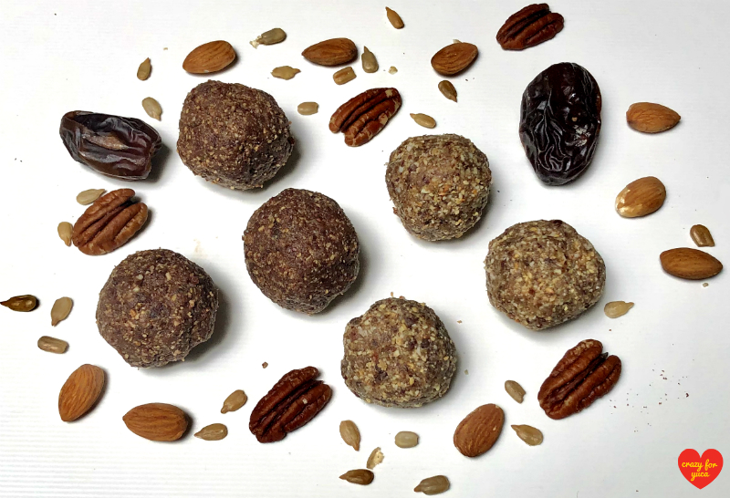 CrazyForYuca.com - Six Paleo No-Bake Date Nut Balls (Cinnamon and Cacao variations) on white plate with scattered nuts and dates. 