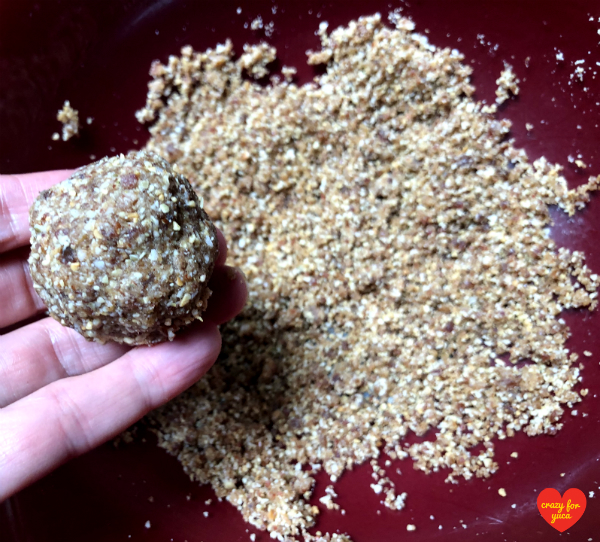 CrazyForYuca.com - A bowl with ground date nut meal in the background with one Paleo No-Bake Date Nut Ball held in hand. Demonstrating proper consistency needed to create balls. 